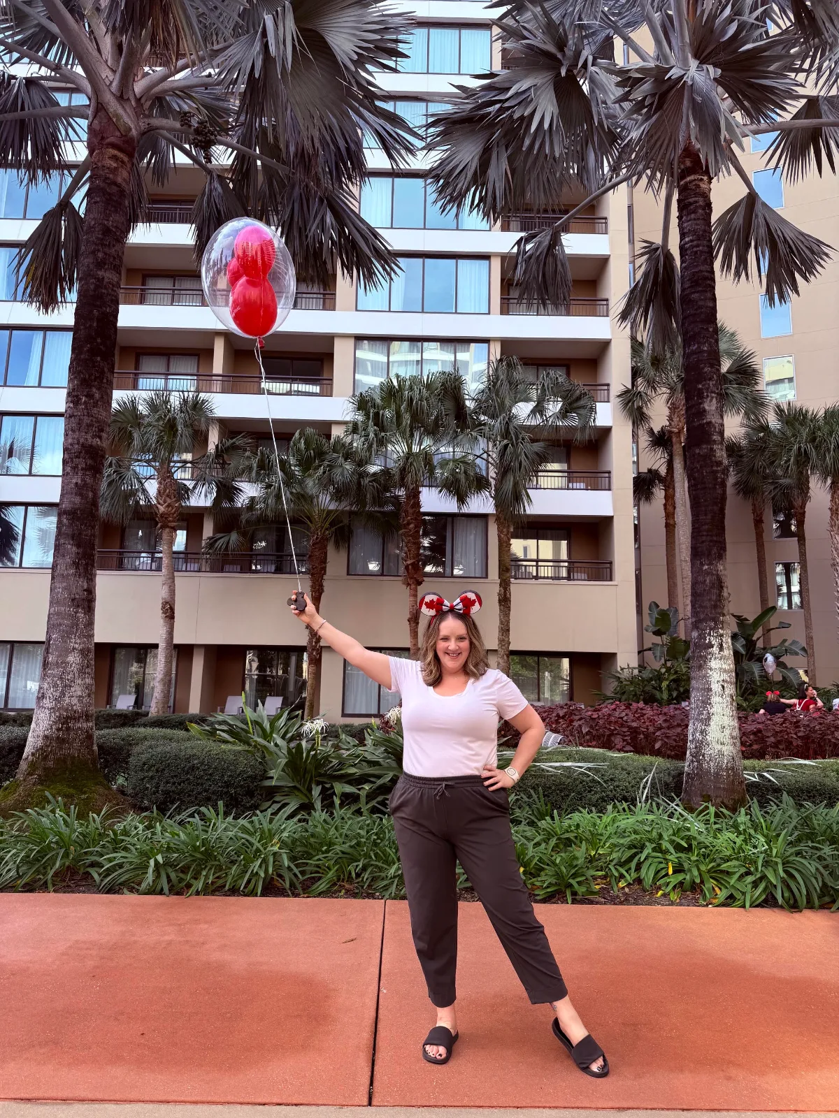 A woman with blonde hair stands in front of a Disney resort hotel holding a red Mickey Mouse balloon. The woman has a Mickey ear headband on, which has Canadian flags on them.